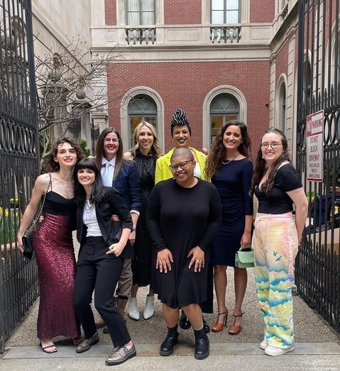 Drew is a white woman with long curly brown hair who is wearing a gorgeous dress with a shimmering burgundy skirt and black sweetheart top. Next to her is Elise who is wearing a pair of black pants, tuxedo top and white shirt. Behind her is Kristen Arnett in blue shirt and tie with blue jacket. Kristen is a tall, white human with long straight brown hair and glasses. Next to Kristen is Riese a tall white woman with blonde hair dressed in black. Next to Riese is Christina, a tall Black woman wearing an electric yellow blazer, black and white hair wrap and makeup. In front of Christina is jourdain, with a shaved head, dressed in black. Next to jourdain is Kayla, a south Asian woman with long wavy brown hair and a navy blue dress. Next to her is Anya a white woman with long brown hair wearing a black top and pants with the sky depicted on them.