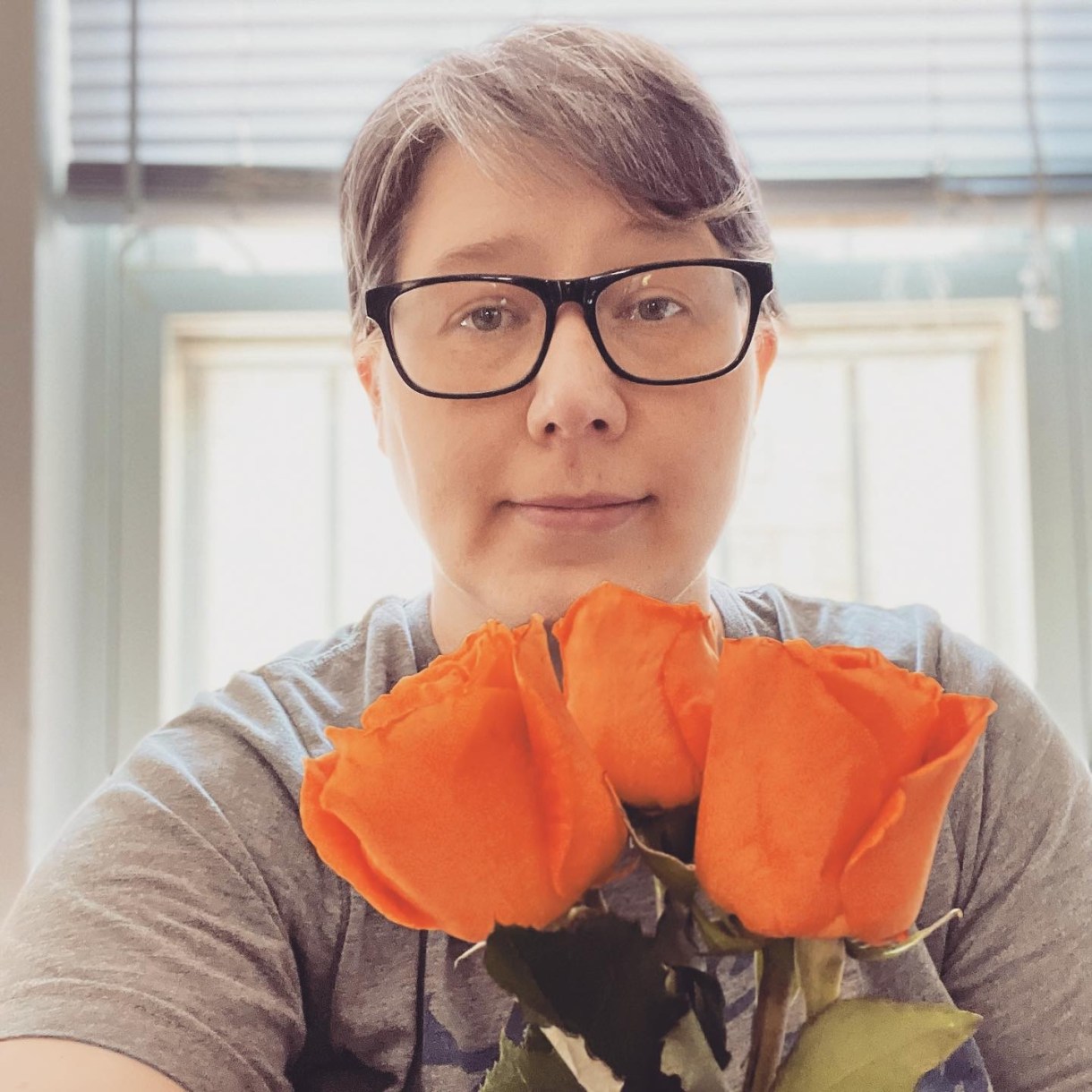 Heather is a white woman with short gray hair and black frame glasses. Here she is holding up three orange roses and raising an eyebrow.