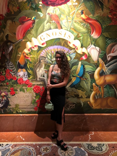 Kayla is a South Asian woman with long wavy brown hair. She is standing in one of her signature outfits with tiny bag in front of an elaborate mural featuring animals and florals.