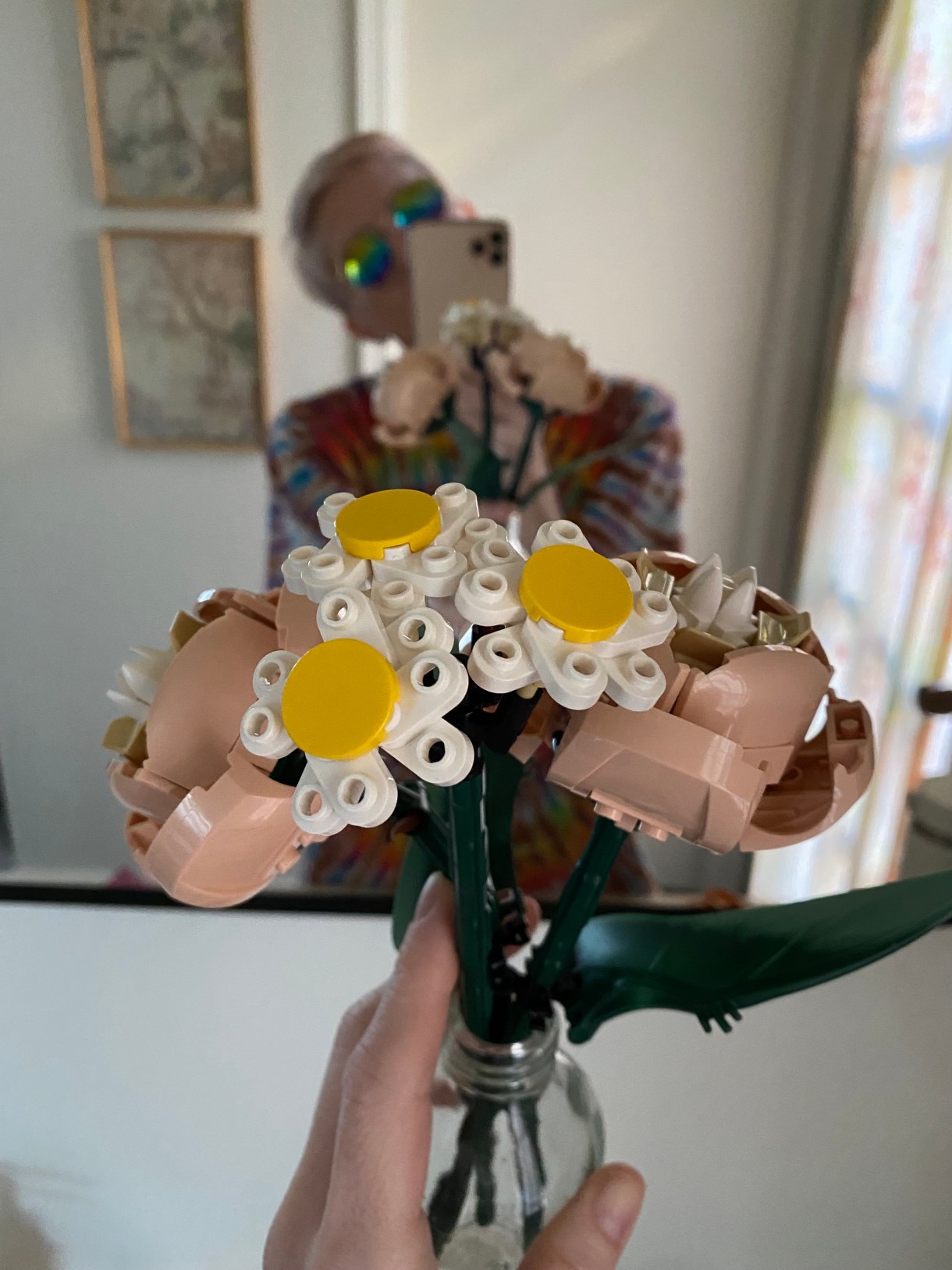 Laneia is a white woman and is holding up a bouquet of lego flowers to a mirror, and is out of focus in the background wearing sunglasses and a tie dye shirt.