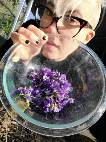 Nicole is a white genderqueer human with bleached white hair, large glasses and short, painted black nails. They are holding a glass bowl of violets that they picked up to the camera.
