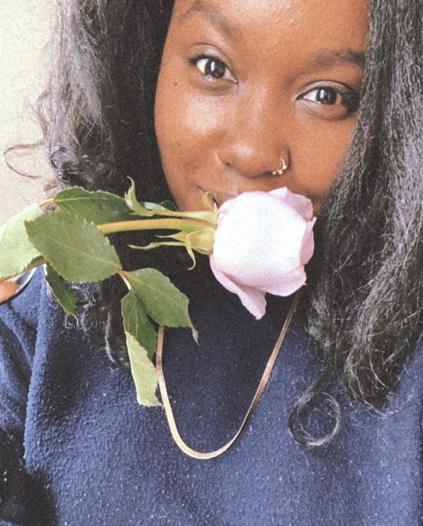 Shelli smiles with a single white rose held up in front of her mouth. Shelli is a Black woman with long black hair and a nose ring.