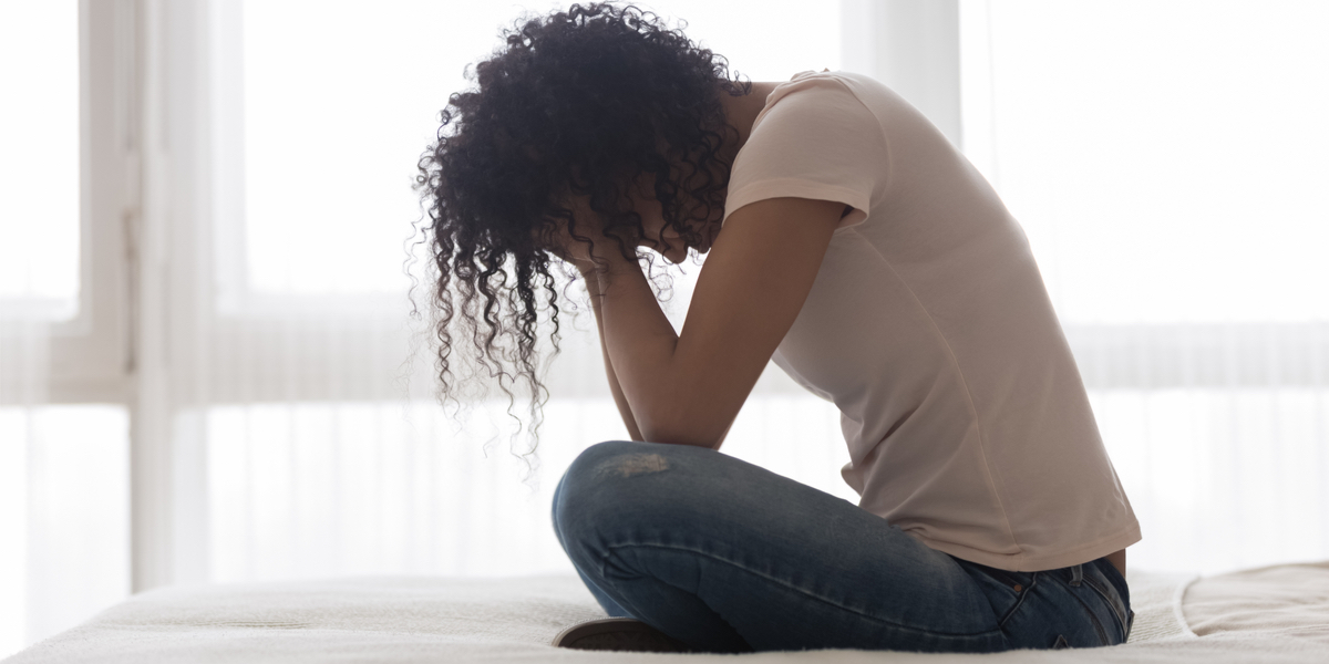 A Black woman with curly hair wearing a pink T-shirt and jeans sits cross-legged on a white bed with her head in her hands. Windows covered in gauzy white curtains are visible in the background.