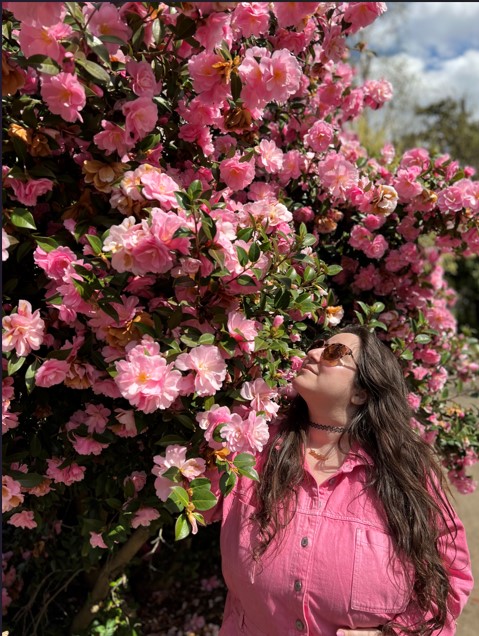 Vanessa, a white woman with long brown hair, is wearing sunglasses and a pink jumpsuit and sniffing a tree covered in pink blossoms