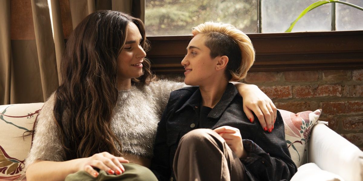 A transfeminine non-binary person and transmasculine gender-nonconforming person look at each other on a couch