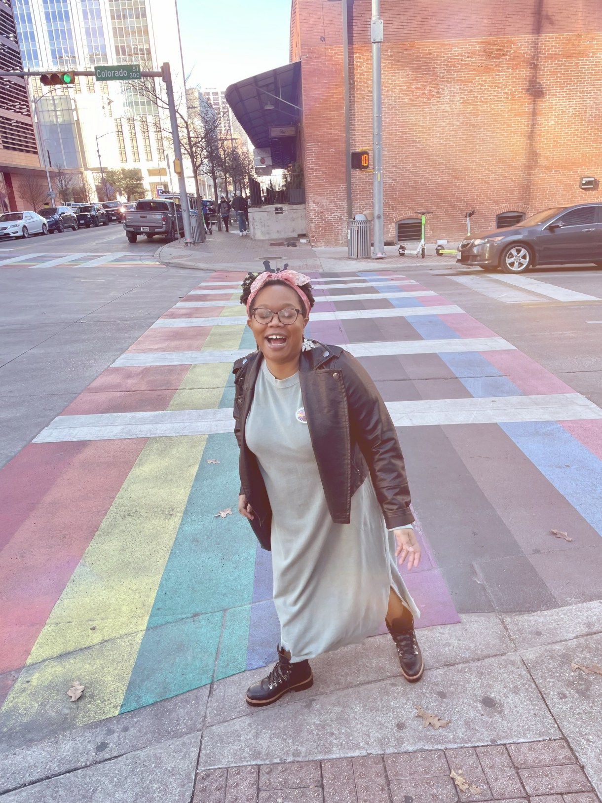 Carmen, a Black woman wearing a dress and jean jacket exclaims joyfully on the edge of a rainbow flag crossing at an intersection.
