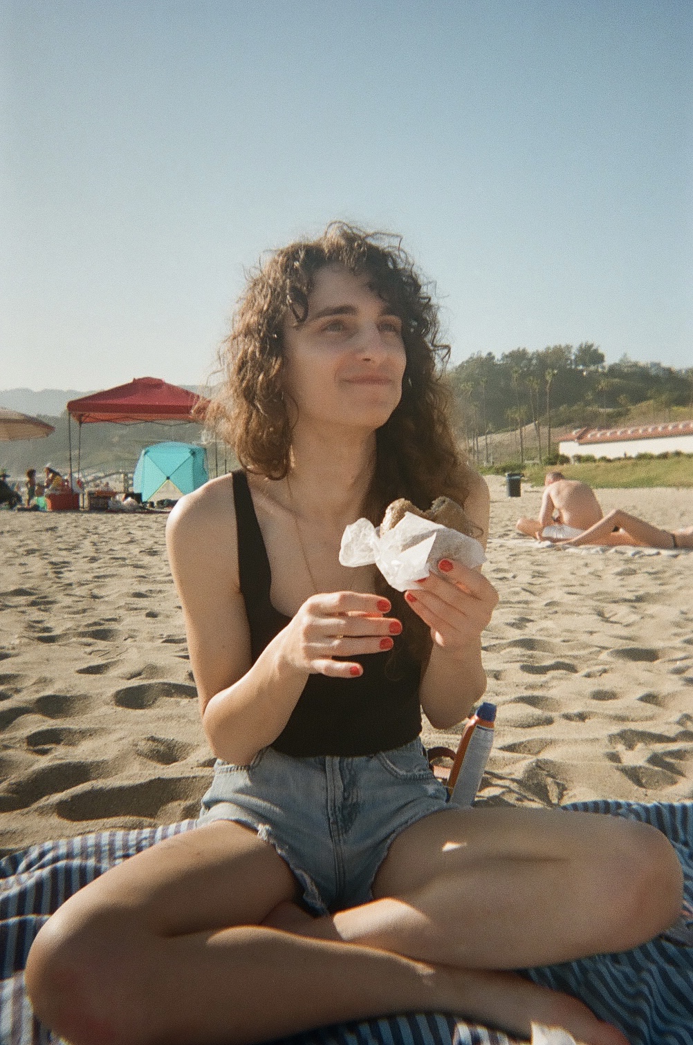 Drew, a white woman with long curly hair, sits cross-legged on a towel on the beach while eating an ice cream sandwich