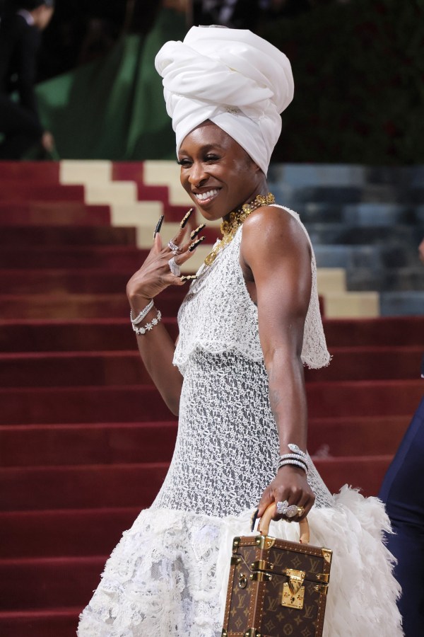 Cynthia Erivo smiles in a white head dress and white lace gown with a small brown purse, she shows off her long nails