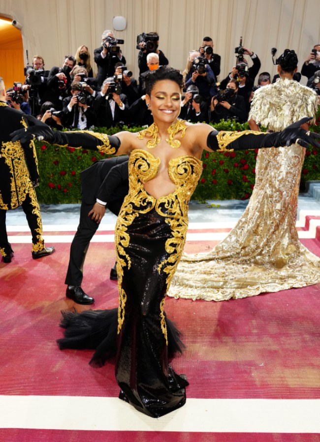 Ariana DeBose in a gold and black structured gown has her arms open wide on the Met red carpet, she's facing directly at camera