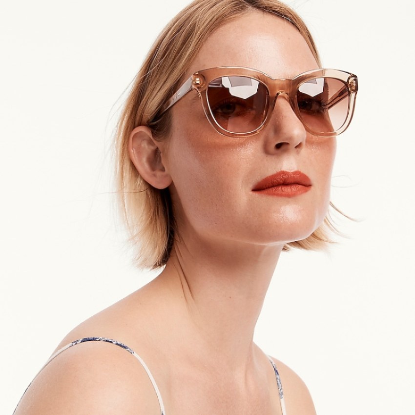 A white femme person with rust colored lipstick and a blonde bob wears oversized square sunglasses with both lenses and frames in a monotone tan color.