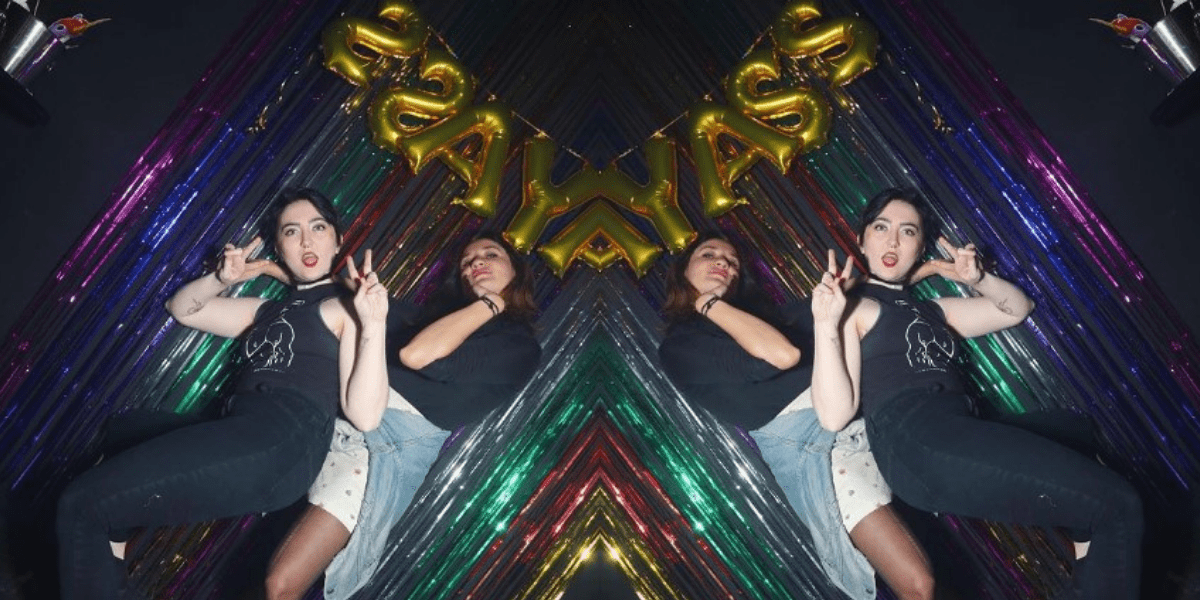 This feature image is made up of one image that is flipped and doubled on the other side, creating a mirror effect. The photo is of Analyssa and Courtney at Gay Asstrology. Analyssa is a a young latinx person with shoulder length brown hair, wearing shorts and a navy shirt with a jean jacket around her waste. She is leaning back against a bright and colorful background of rainbow streamers. Courtney is a white woman, and in front with short black hair, wearing all black with red lipstick, and giving double V for Victory signs with her fingers.