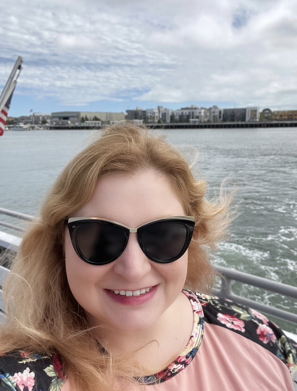 Katie, a white person with gingery hair, is wearing sunglesses and appears to be on a boat, out on the water on a sunny day.