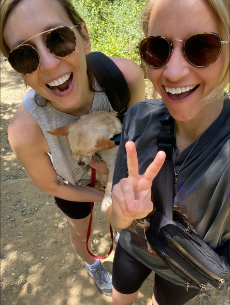 Riese, a white woman wearing sunglesses, poses with a friend and carol the dog while on what looks like a hike outside