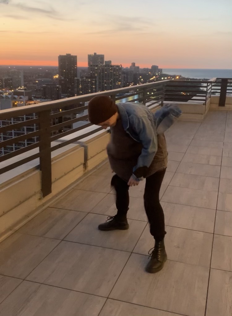 Ro, a white human, is wearing a flannel shirt, beanie, and black boots and appears to be dealing with some kind of wind situation on a city rooftop.