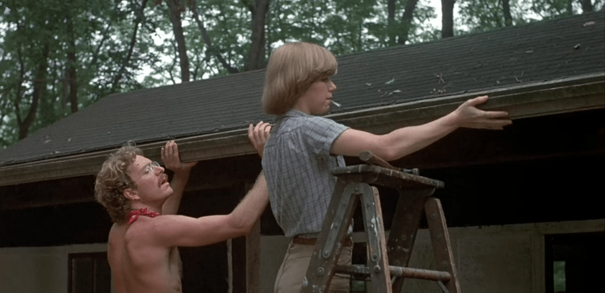 A shirtless white man and a white woman in khakis, a short sleeve buttondown, and a bowl cut work on a cabin roof in Friday the 13th