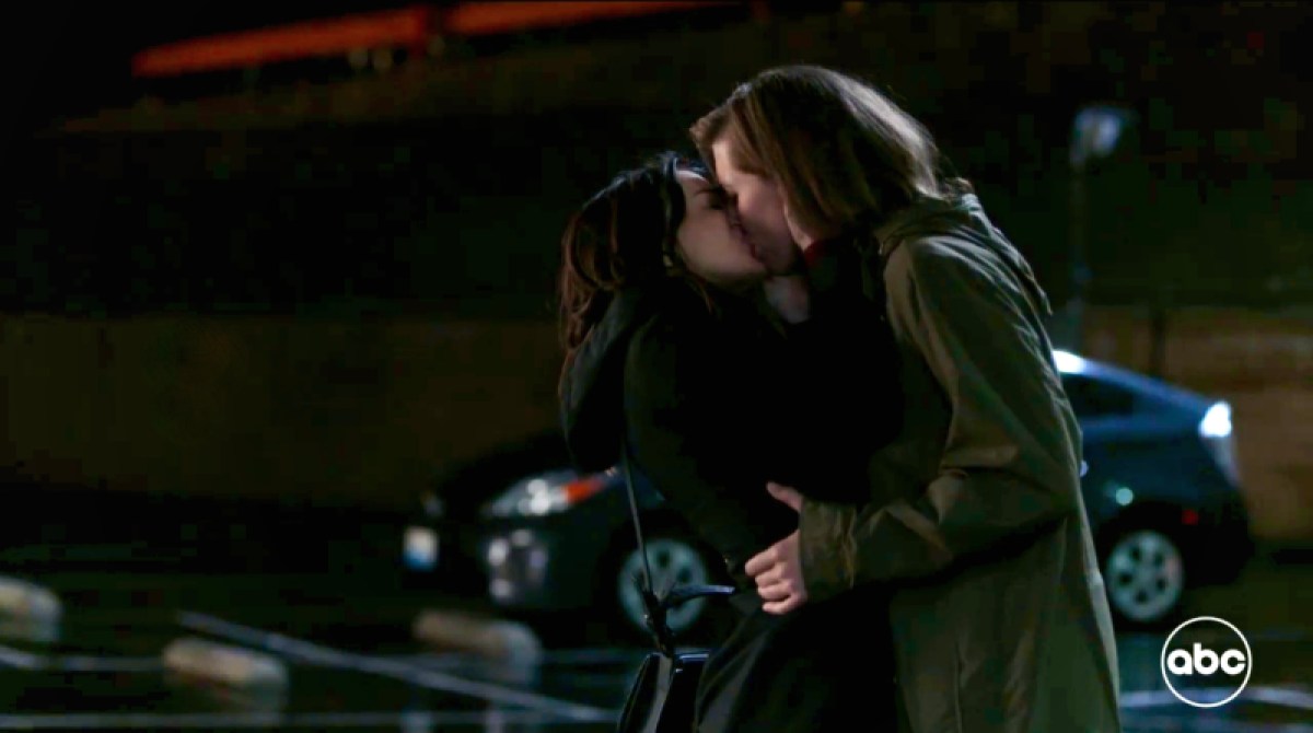 In a still from Grey's Anatomy, in a dark parking lot at nightfall, Amelia and Kai kiss. Kai's hands are on Amelia's waist.