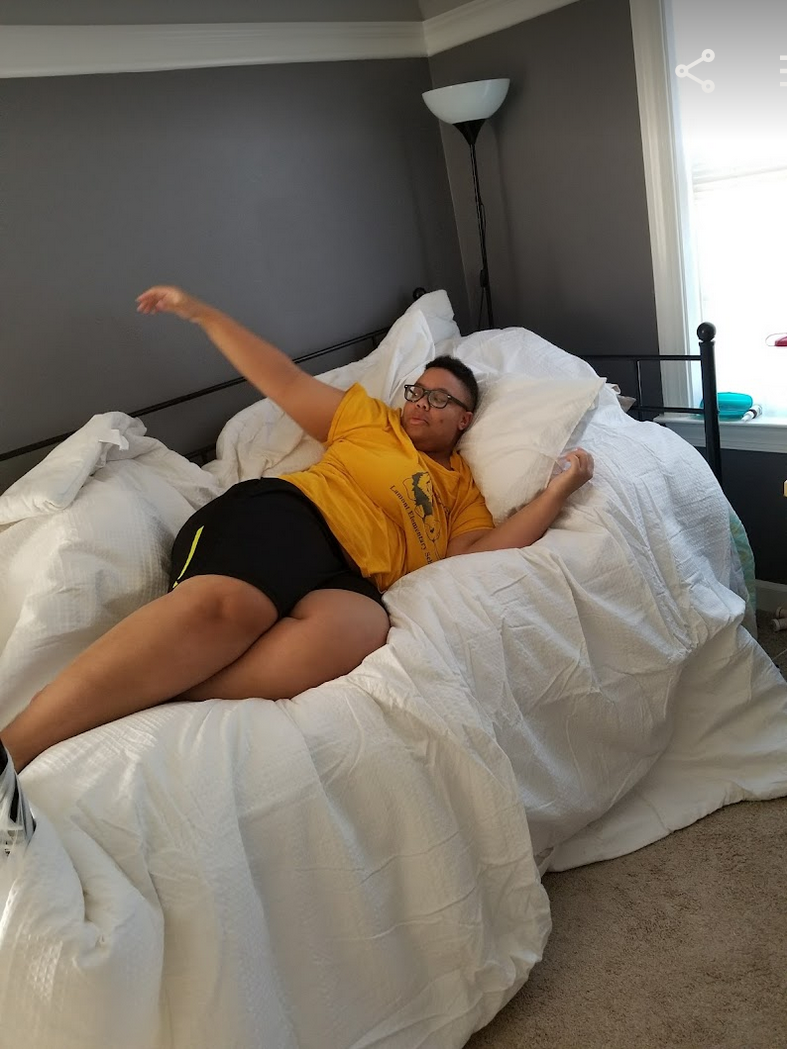 A.Tony Jerome lounges on a day bed on top of a white cover. They are a Black human wearing glasses, shorts and a yellow shirt, and they have short hair. They look like they are in the middle of gesturing or saying something.