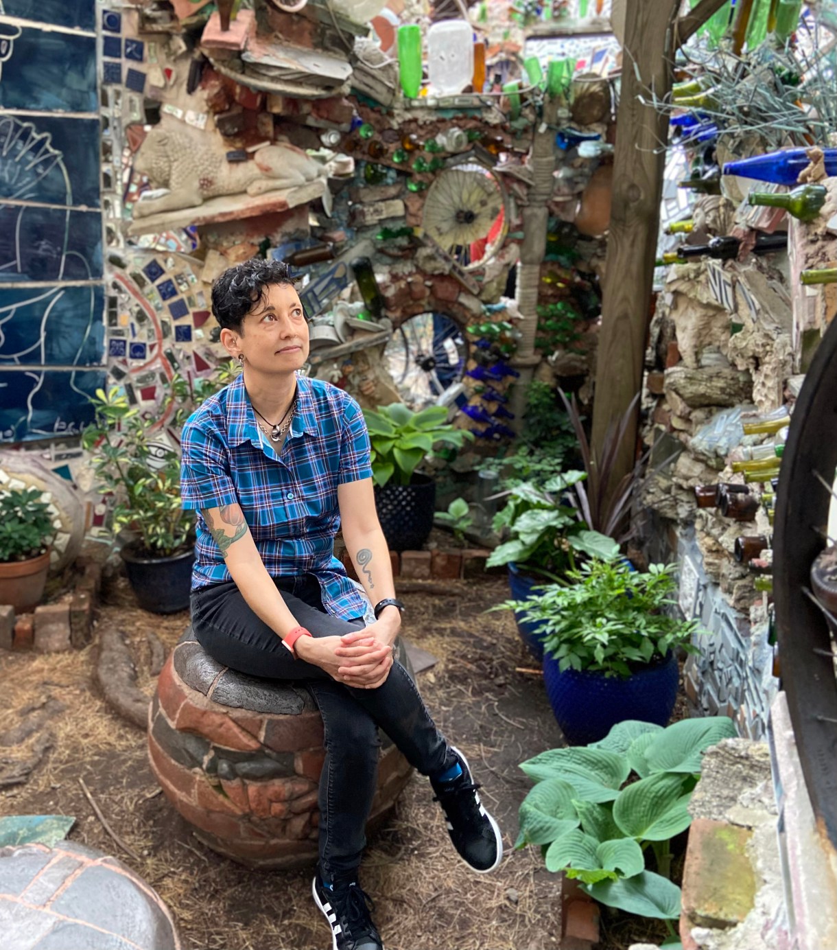 Tracy an Asian woman sits,  with short hair, sits with legs crossed on a stool or bench situation in front of a bunch of mosaics
