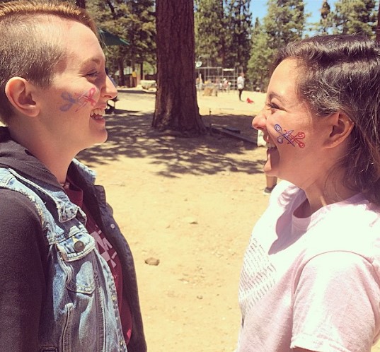 Courtney is white, with blond hair that is shaved underneath and floppy on top. Analyssa is Latinx with brown hair the length of her shoulders. In this photo they are looking at each other and smiling with matching scissoring face painting art on their cheeks. The face painting looks like the Autostraddle scissoring design.