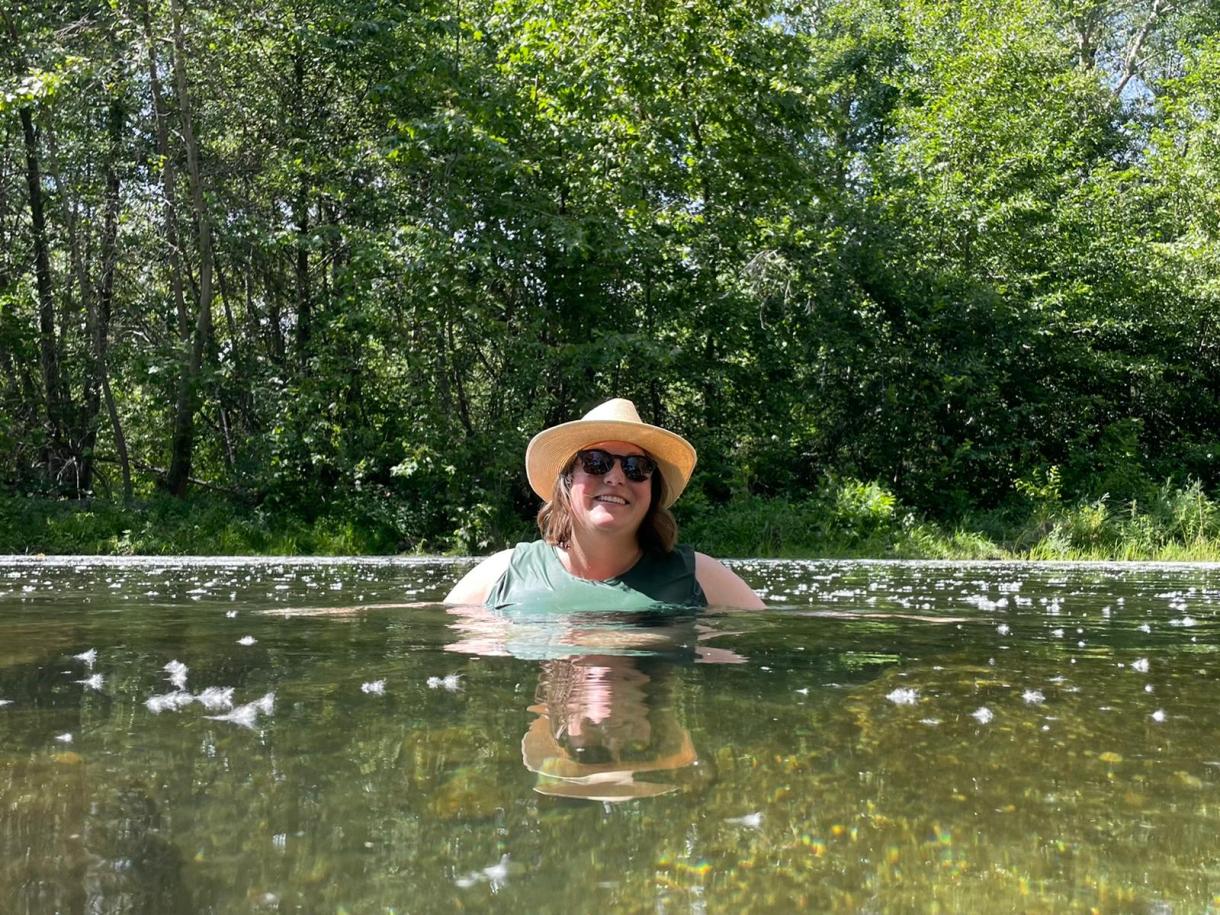 Darcy, a white human, smiles while wearing a hat and sunglasses, submerged in some kind of pool near a bunch of trees