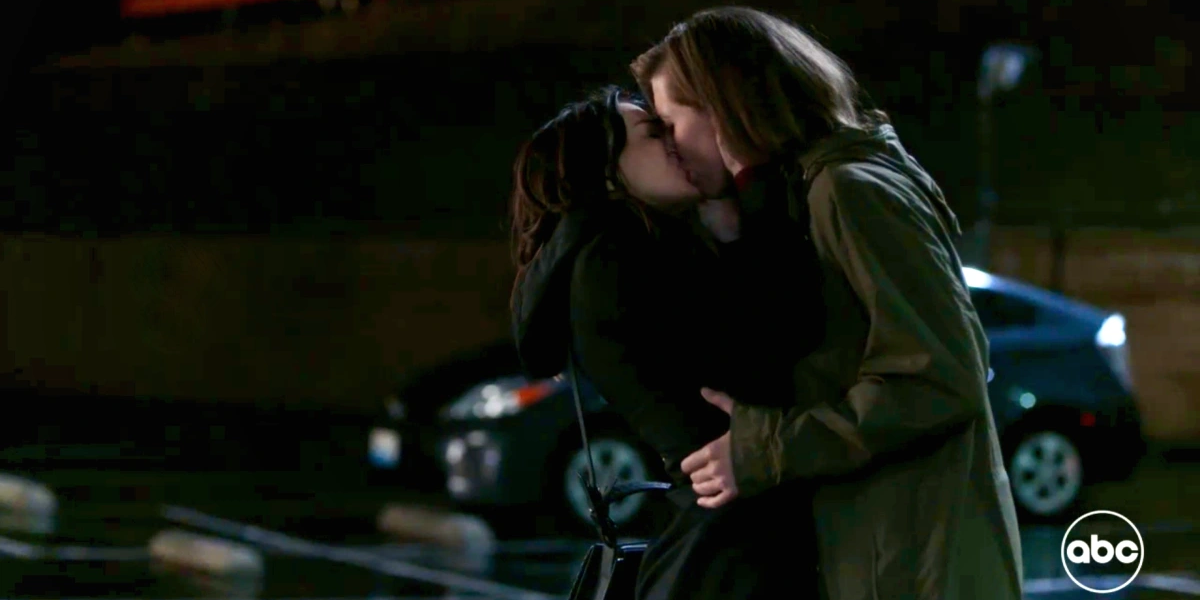 In a still from Grey's Anatomy, in a dark parking lot at nightfall, Amelia and Kai kiss. Kai's hands are on Amelia's waist.