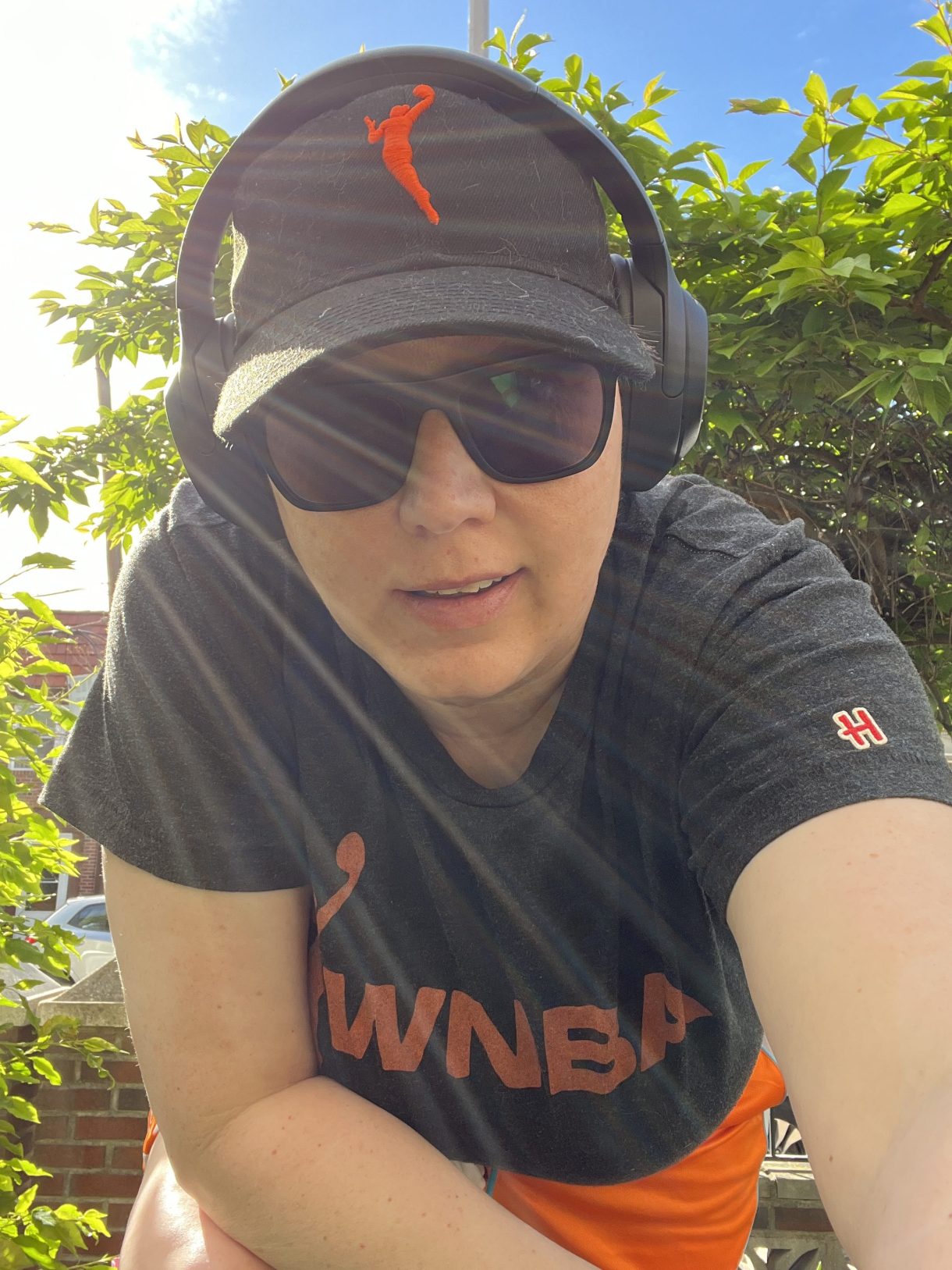Heather, a soft butch white woman leans forward in sunglasses, headphones, and full WNBA regalia from hat to tee shirt to shorts.
