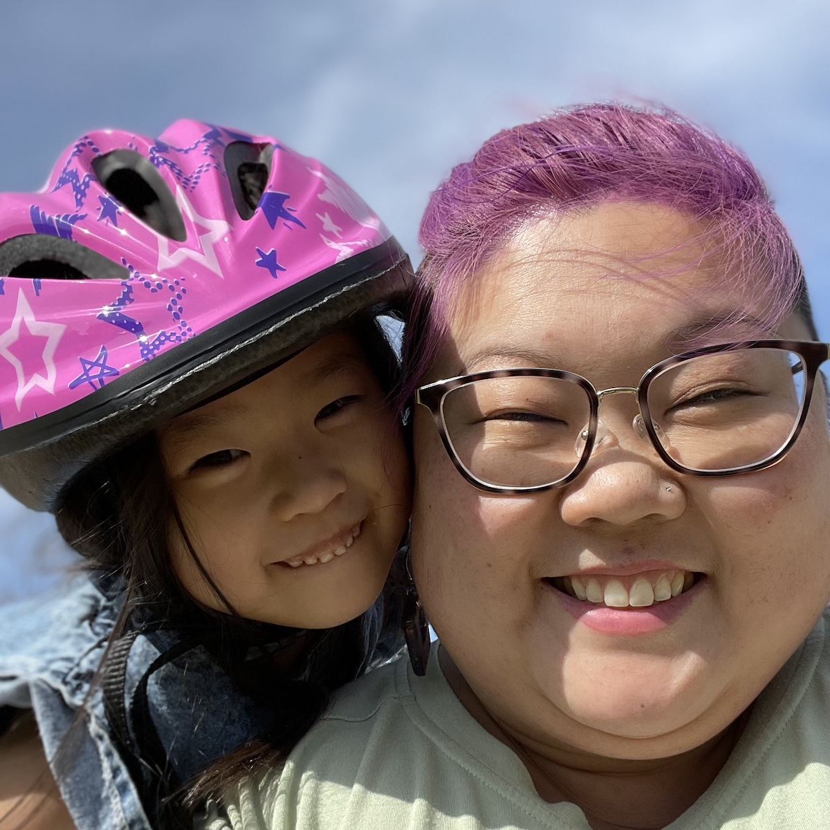 KaeLyn, an Asian woman with bright purple hair, smiles at the camera as her kid leans over her shoulder and smiles while wearing a bright pink bicycle helmet.
