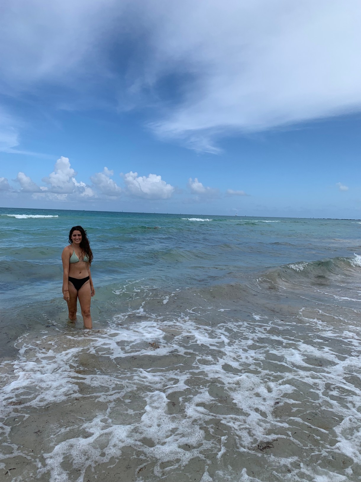 Kayla, a South Asian woman with long brown hair, stands in the ocean, wearing a bathing suit, on a semi-clowdy day.