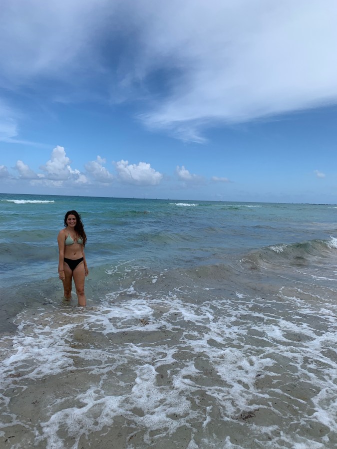 Kayla, a South Asian woman with long brown hair, stands in the ocean, wearing a bathing suit, on a semi-clowdy day.