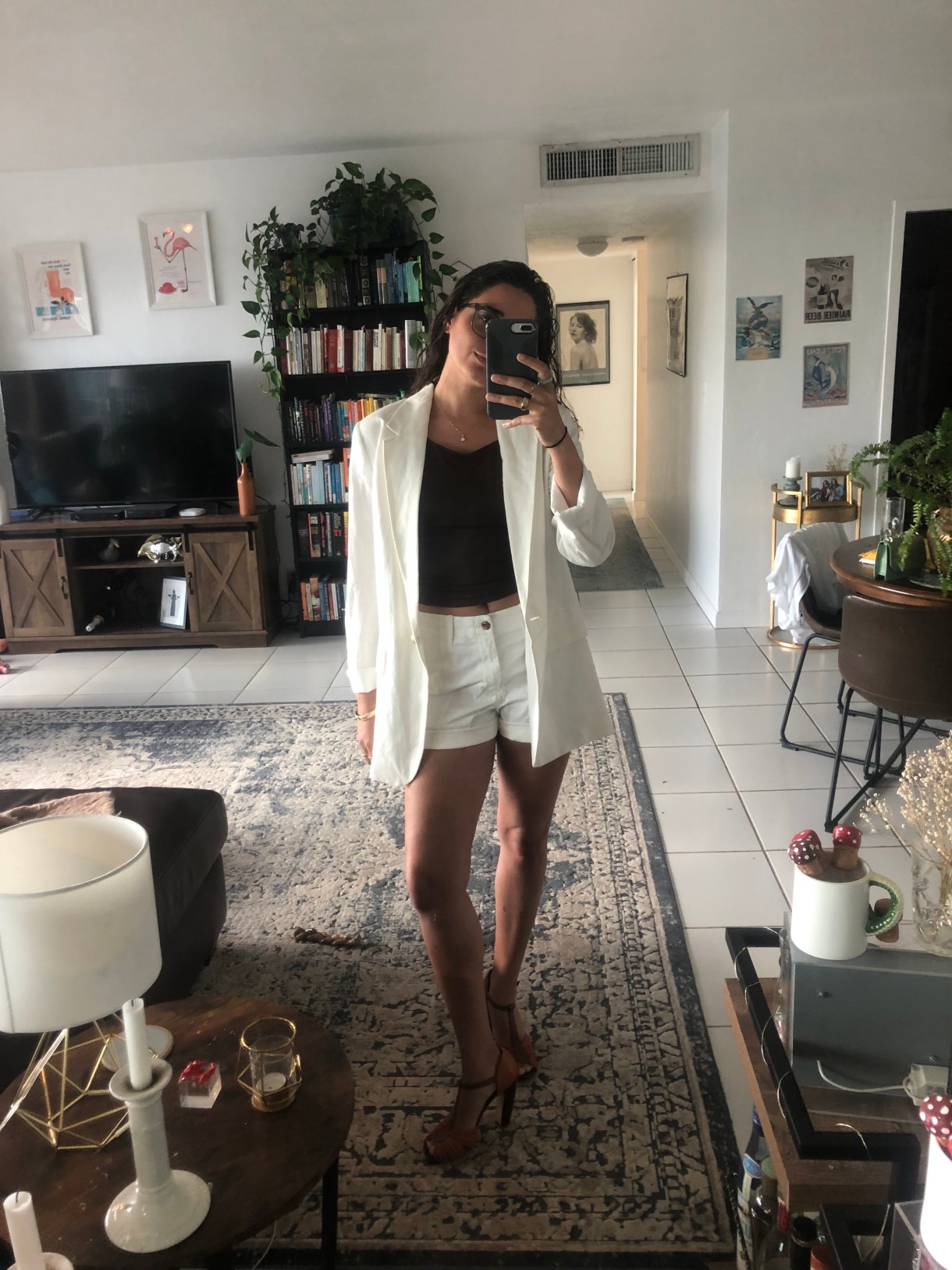 Kayla Kumari Upadhyaya wears a long white blazer over a brown cami and white shorts. She is also wearing high brown heels and taking a mirror selfie in her living room.