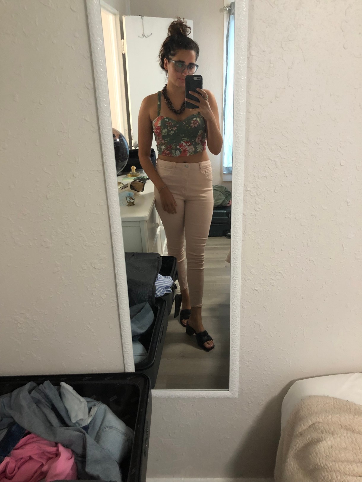 Kayla Kumari Upadhyaya has her hair in a bun and is wearing a floral bustier crop top, light pink pants, black heeled mules, and a brown chunky wooden necklace. She is taking a mirror selfie in an Airbnb bedroom.
