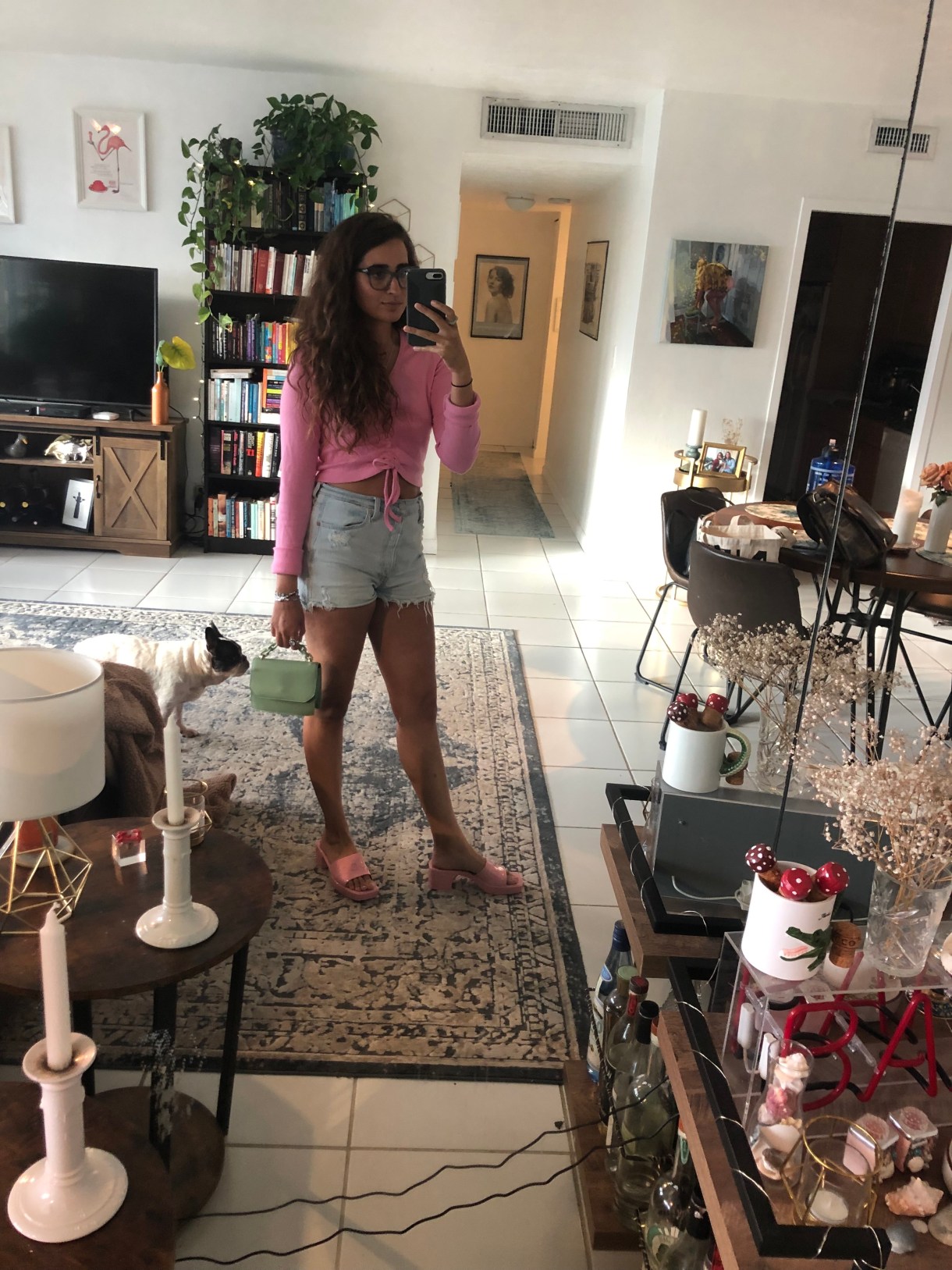 Kayla Kumari Upadhyaya has her hair down and is wearing a long sleeved pink crop top that ties in the middle and cut-off jean shorts and pink heeled slides. She is holding a tiny green purse and taking a mirror selfie in her living room. Lola, a black and white French bulldog, is in the background.