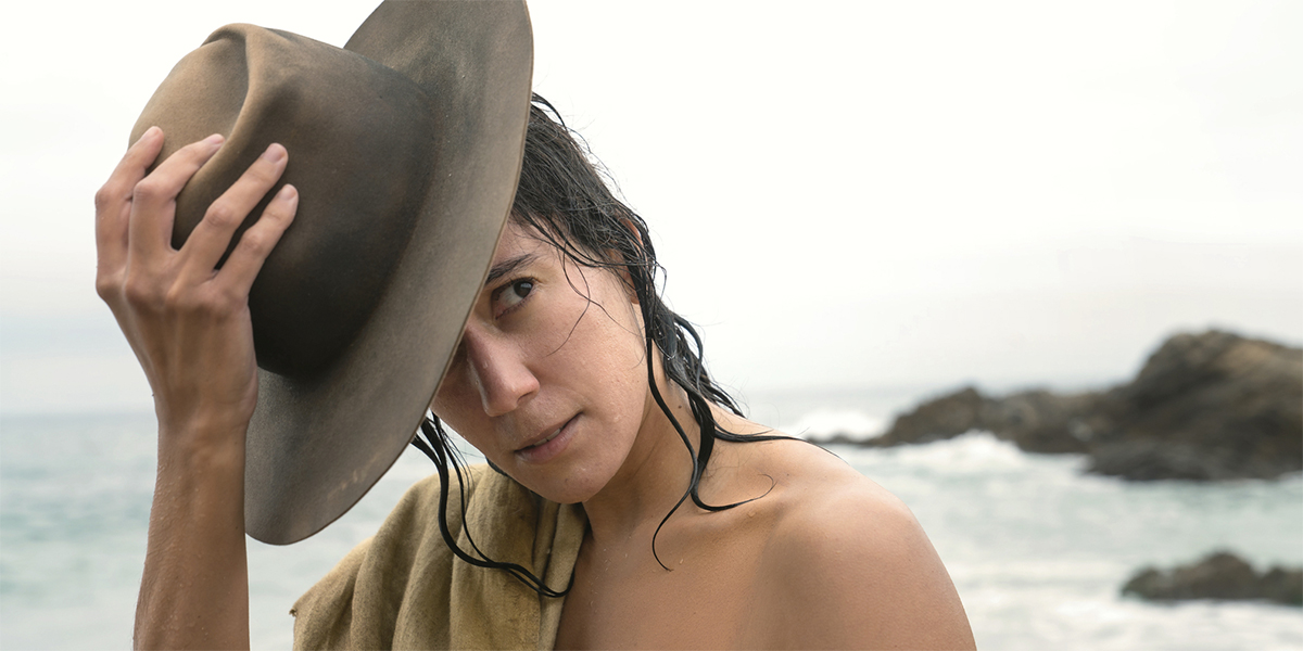 In Our Flag Means Death, Vico Ortiz takes off their hat while standing shirtless in front of the ocean