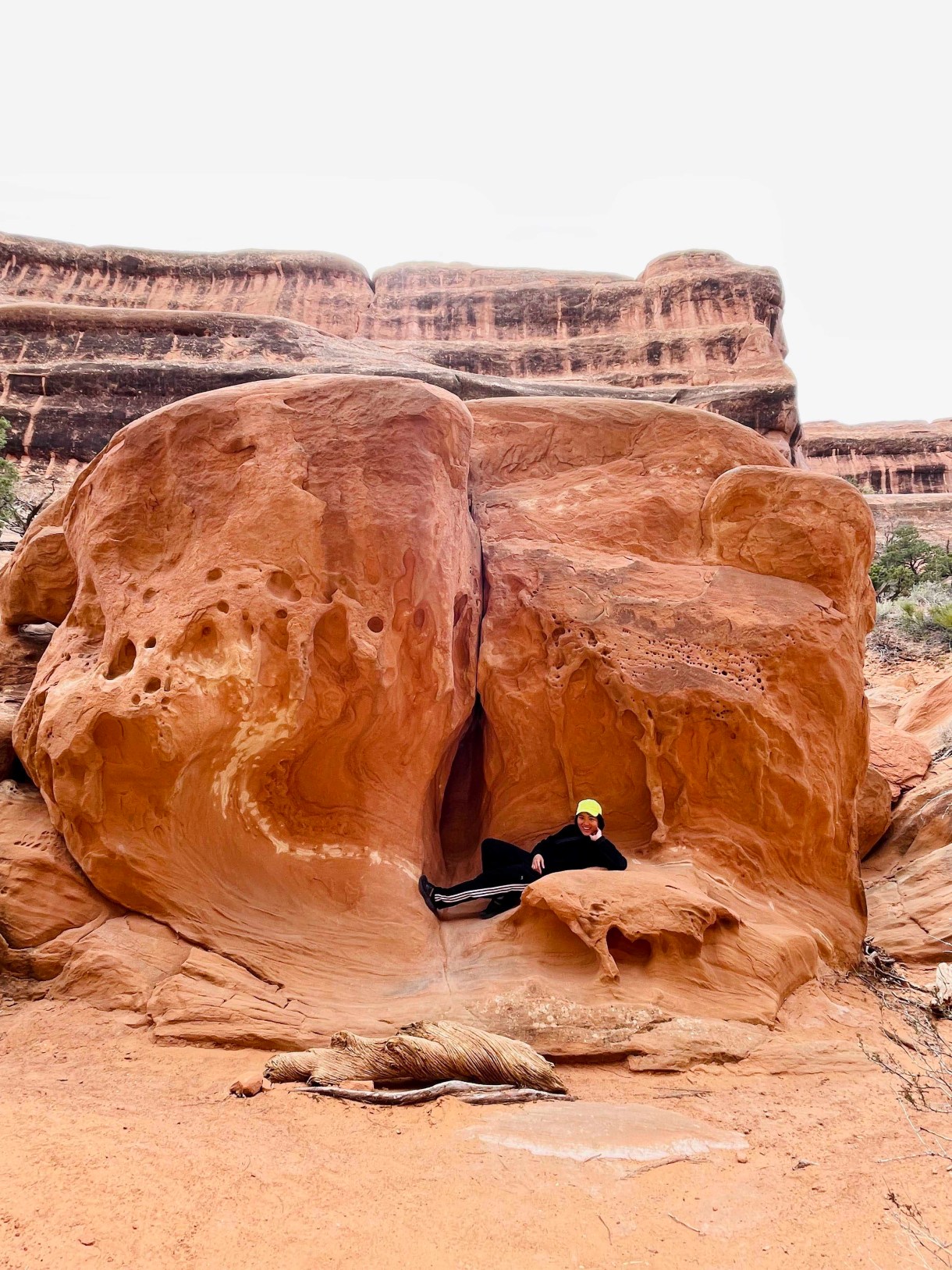 Viv, an Asian human wearing athletic clothing and a baseball cap, poses on their side in front of a Georgia O'Keefe sculpture made out of a giant rock. It looks like a vagina.