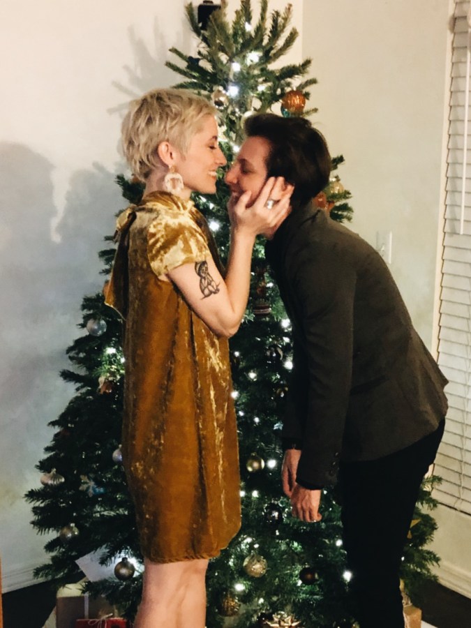Laneia and Amanda pose in front of a Christmas tree. Laneia is holding Amanda's face in her hands. They're both smiling hugely. Laneia is wearing a golden velvet babydoll dress. Amanda is wearing a deep brown suit jacket and black slacks.