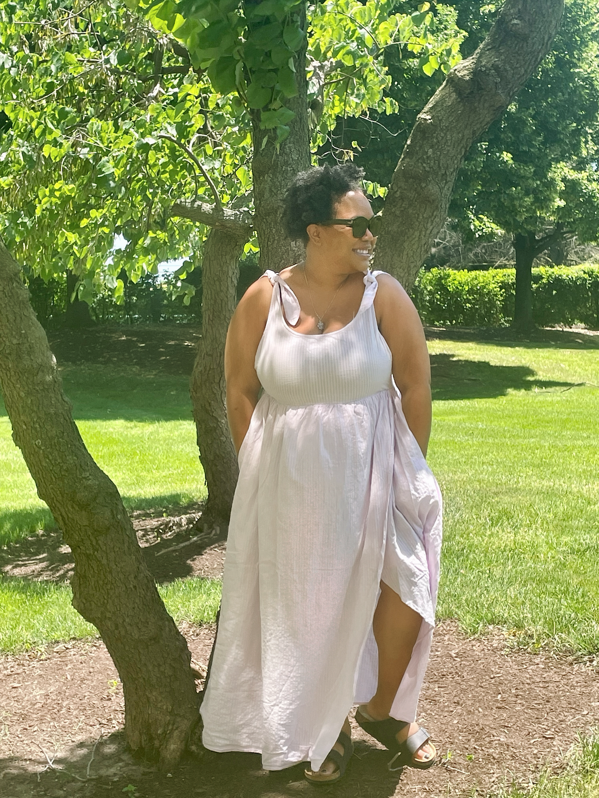 Carmen looks like an absolute goddess in this white or light pink full length sundress. She is a Black woman wearing sunglasses with medium-short curly black hair. Carmen is wearing Birkenstocks and smiling in front of a grove of trees.