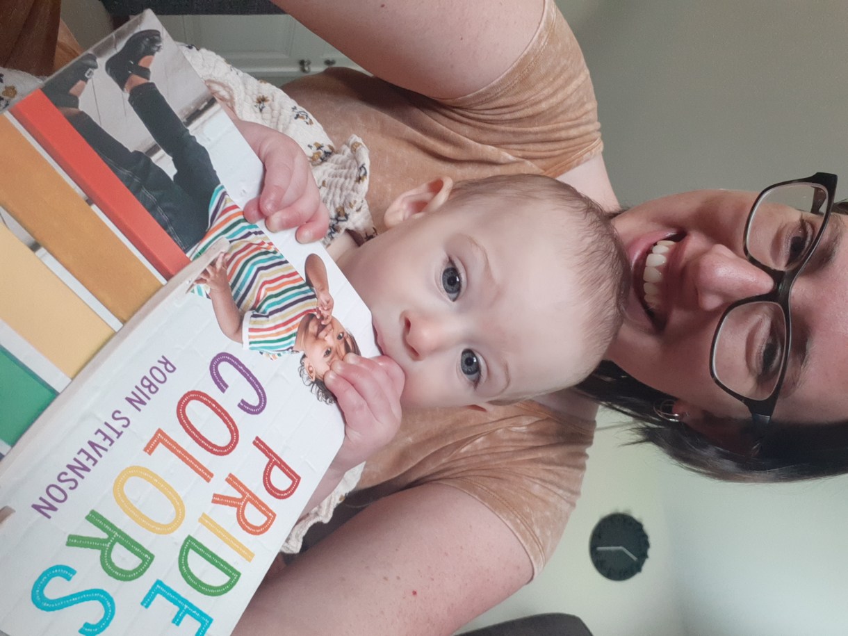 Casey is here with her baby who is chewing on a "pride colors" toy. Casey is a white woman with medium short brown hair and glasses. She is smiling and her baby is concentrating on the book.