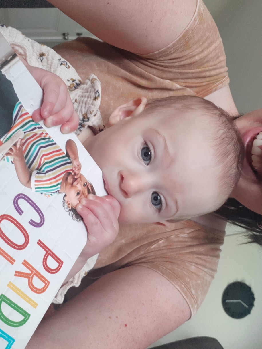 Casey is here with her baby who is chewing on a "pride colors" toy. Casey is a white woman with medium short brown hair and glasses. She is smiling and her baby is concentrating on the book.