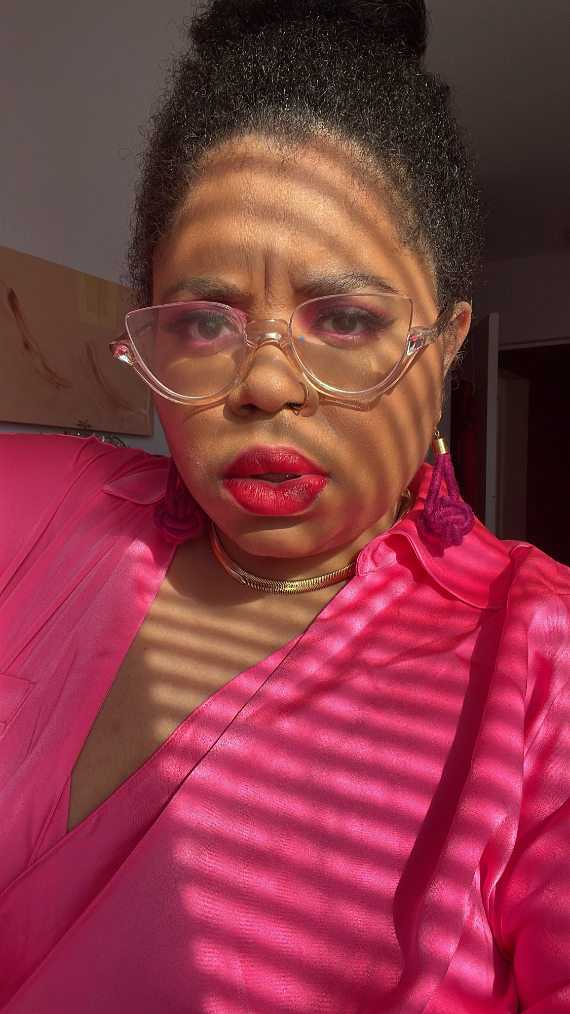 Dani is here in a pink outfit with matching eyeshadow, lipstick and glasses! Dani is a Black woman, her hair is up in a bun and she is wearing a golden necklace. Dappled sunlight from blinds washes over her.