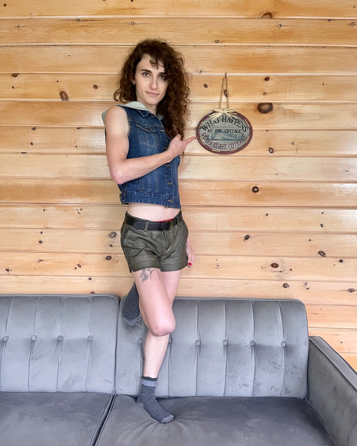 Drew is standing on one foot on a couch in what is presumably a cabin, pointing to a sign that reads 'whatever happens in this cabin, stays in this cabin.; Drew is a white woman with curly, long brown hair wearing short olive shorts and a denim vest.