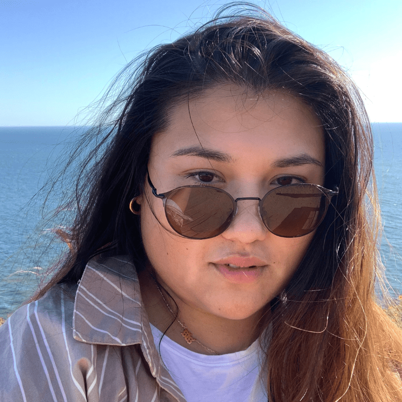Em is a mixed race Southeast Asian person with round dark sunglasses, their hair swept to the side, in front of water