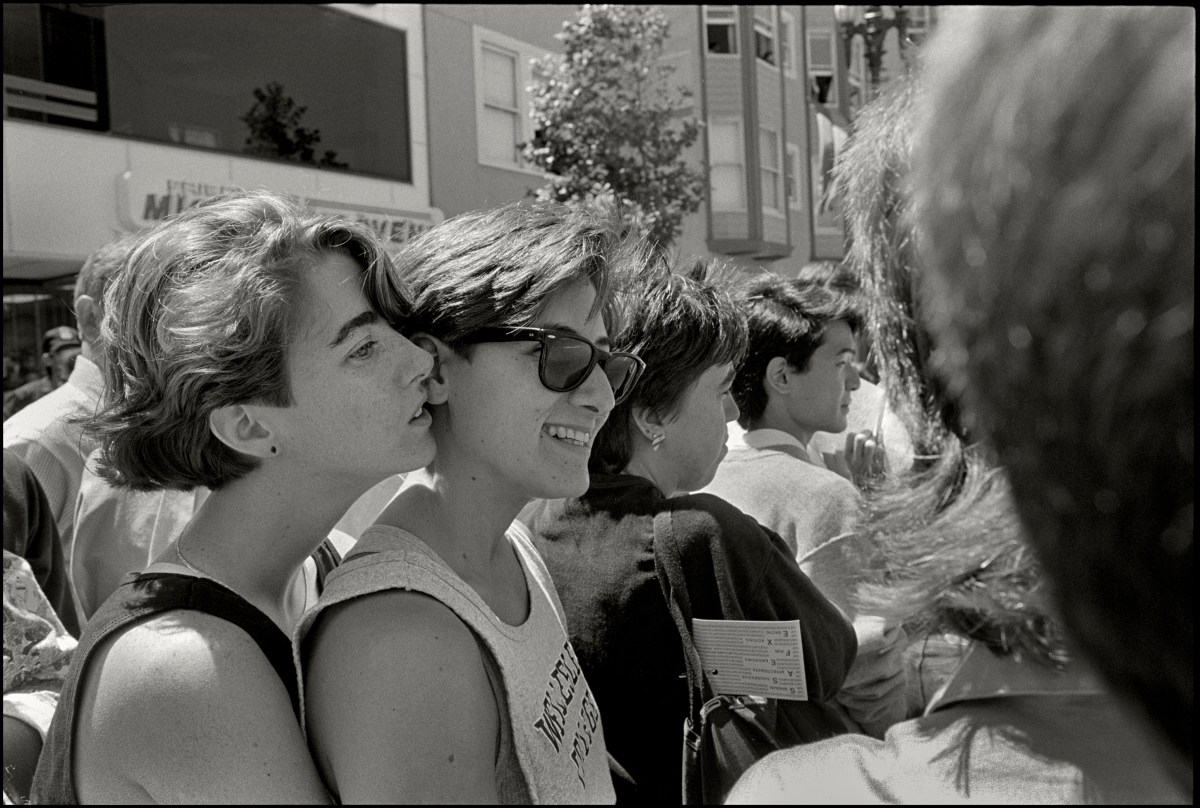 A black and white photo of two lesbians embracing, one is holding the other from behind. They both have short brunette hair.