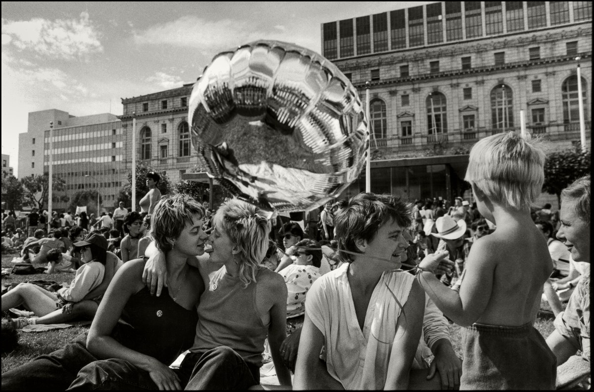 A large group of people sit on the grass but in the center is two lesbians with mullets with their faces close together, one sticking out her tongue at the other.