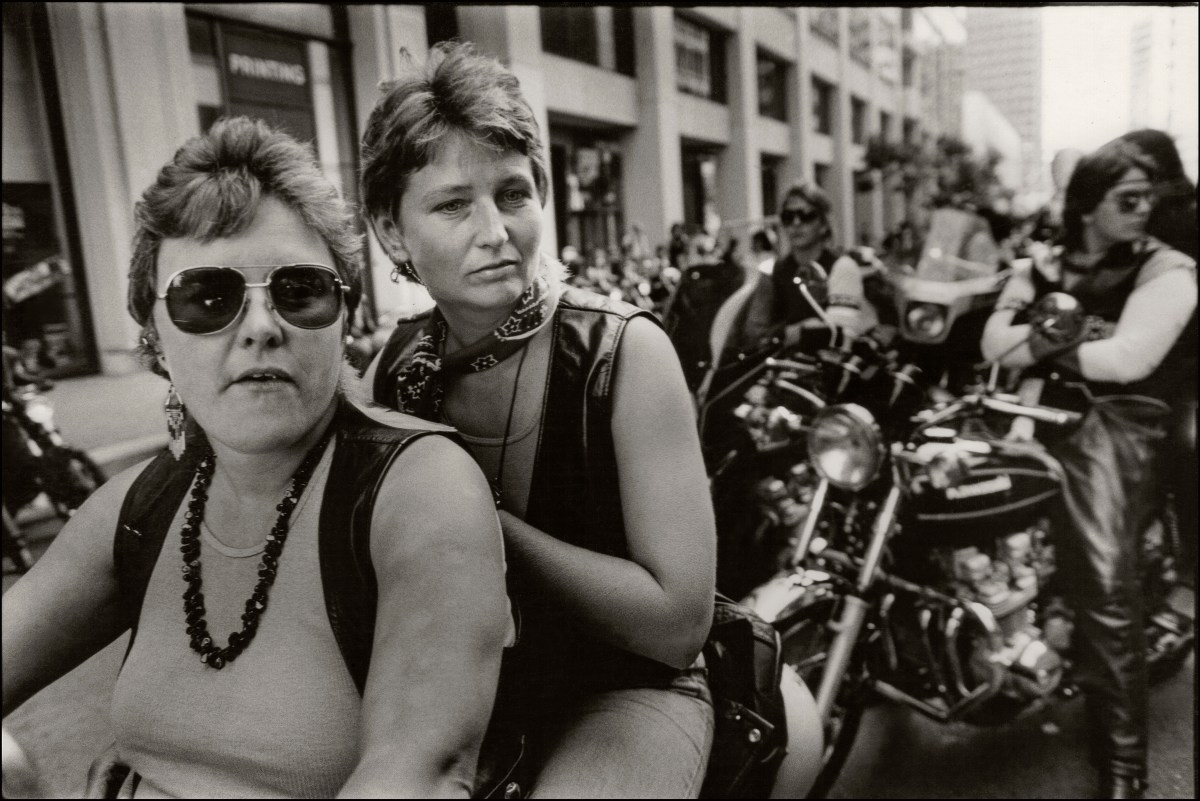 A close up portrait of two women at the dykes on bikes parade, they share a motorcycle and the first woman is fat wearing aviator sunglasses. The second woman is thin with a leather vest.