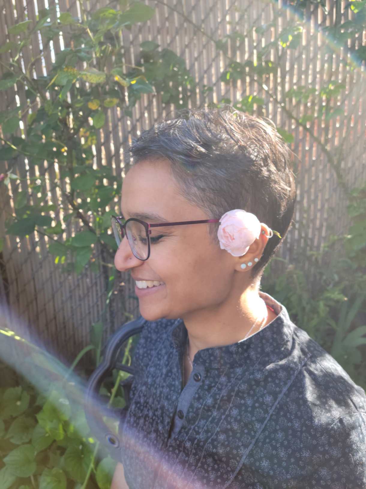 Himani is smiling in sunlight in front of a trellis covered in vines, with a light pink or write rose tucked above her ear. She is a south Asian woman with short dark salt and pepper hair, glasses, and several ear piercings. She is wearing a pattenred shirt.