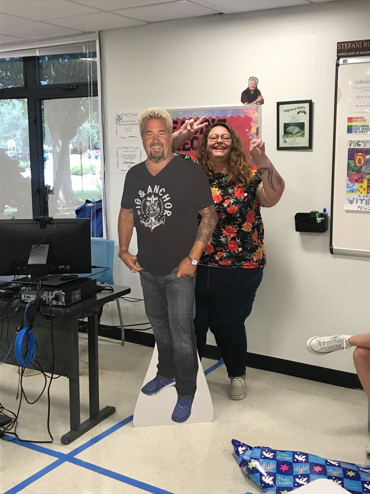 Here Stef stands behind a cardboard cutout of Guy Fieri holding up the V for victory sign with each hand. Stef is wearing a floral print shirt, jeans, sneakers and glasses and is in a classroom. A tiny cutout of guy fieri also peeks out from atop a bulletin board.