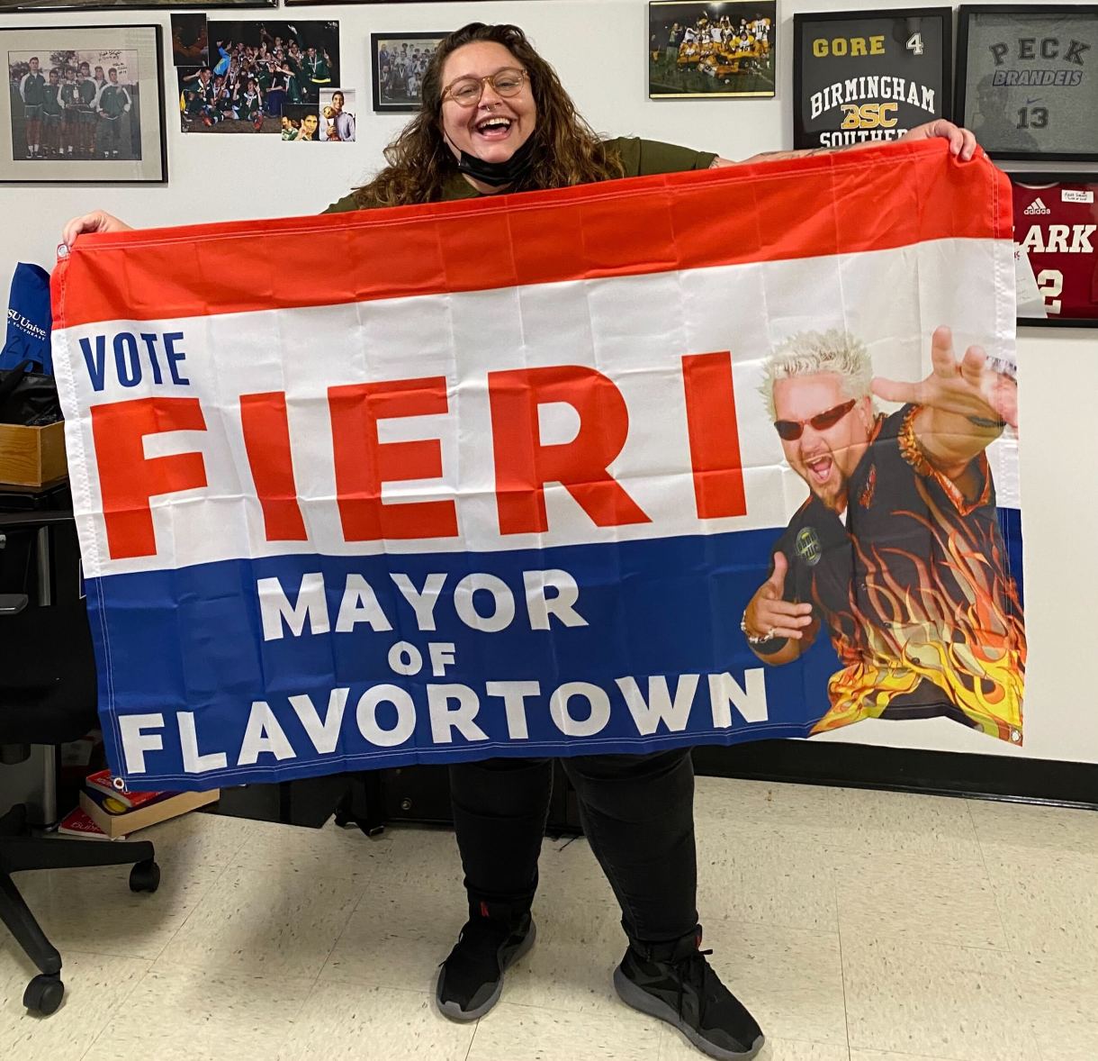 Stef stands, overjoyed, unfurling a giant flag in front of them. The flag is a Vote Fiere Mayor of Flavortown flag with huge, bold, text in red white and blue. Guy Fieri is on it in his signature exuberant pose, bleached hair and firey shirt. Stef is a white person with glasses and long brown hair who in this photo is largely obscured by this huge flag.