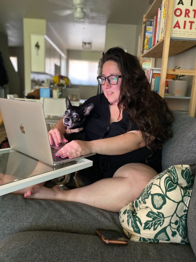 Vanessa, a white woman with long brown hair and glasses, sits on a comfy gray couch, a kind of worktable above her, and her tiny dog Zucchinni in her lap. she is wearing either a black romber or a black two piece shirt and shorts situation.