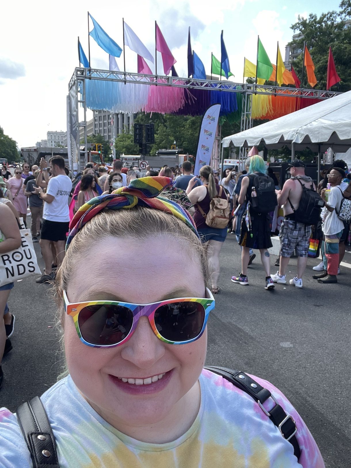 Katie is at a Pride event! Katie is a white woman wearing rainbow sunglasses and a rainbow headband tied at the top of her blonde hair. You can vaguely see people marching in the background.