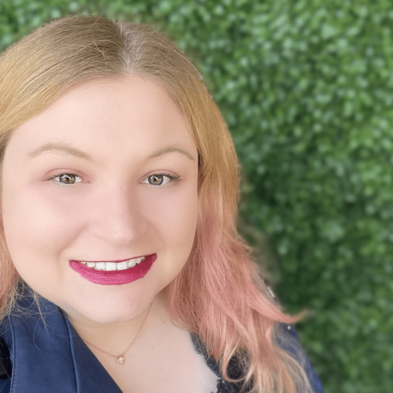Katie is a white woman with blonde hair and ombre pink highlights, she has pink lipstick and is in front of a green bush wall.